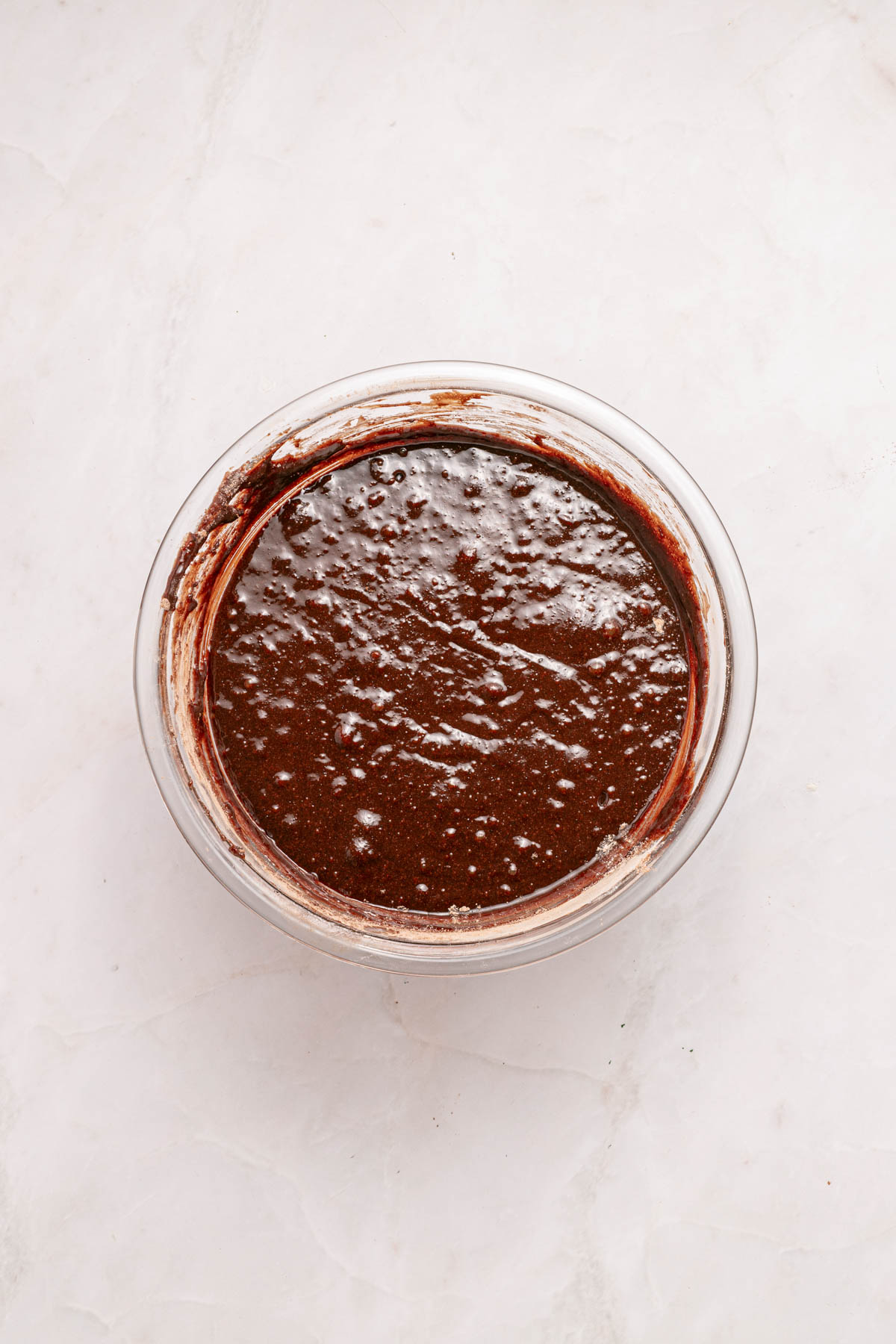 A glass bowl filled with chocolate cake batter sits on a light-colored marble surface.