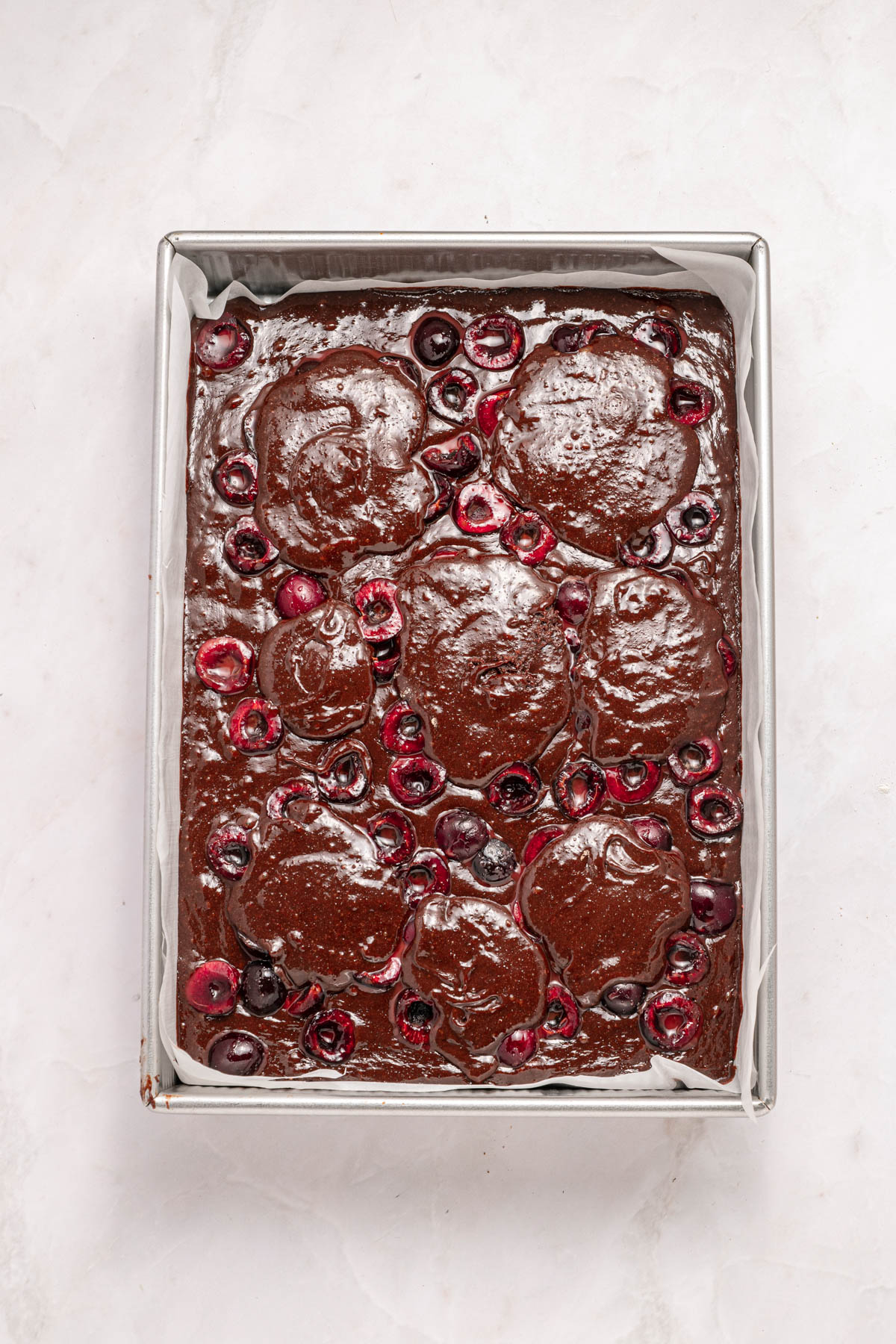 A rectangular pan filled with unbaked chocolate batter and halved cherries on top, placed on a white surface.