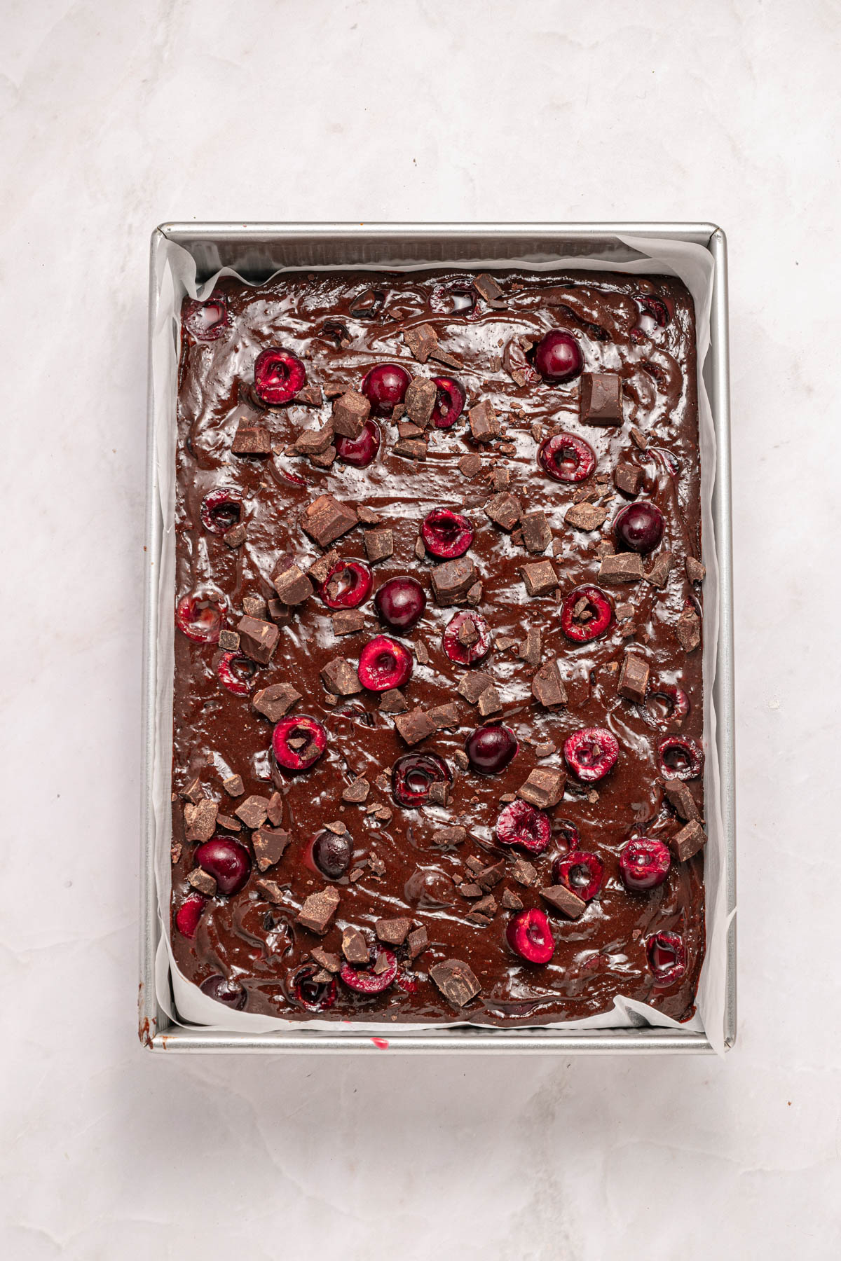 A rectangular baking pan filled with unbaked chocolate batter, topped with halved cherries and chunks of chocolate, on a light surface.