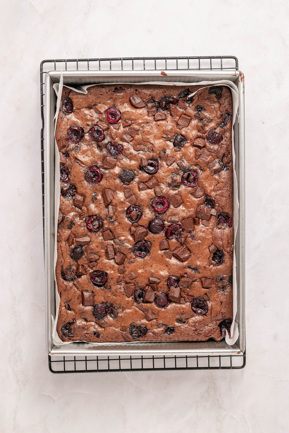 A rectangular tray of baked chocolate cherry brownies on a wire cooling rack, lined with parchment paper.