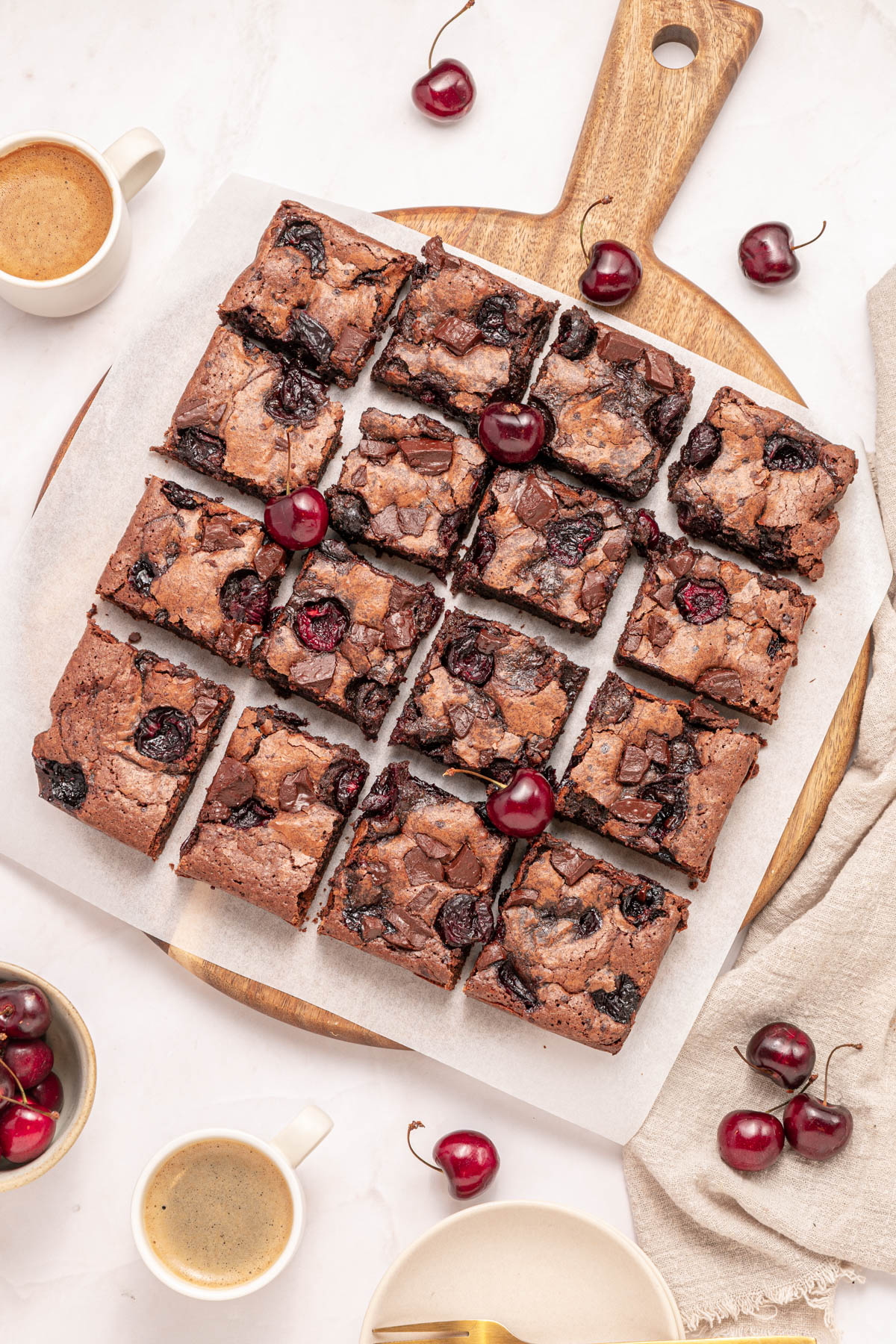 A batch of cherry brownies cut into squares on parchment paper, garnished with fresh cherries, surrounded by cups of coffee and bowls of cherries on a light table.
