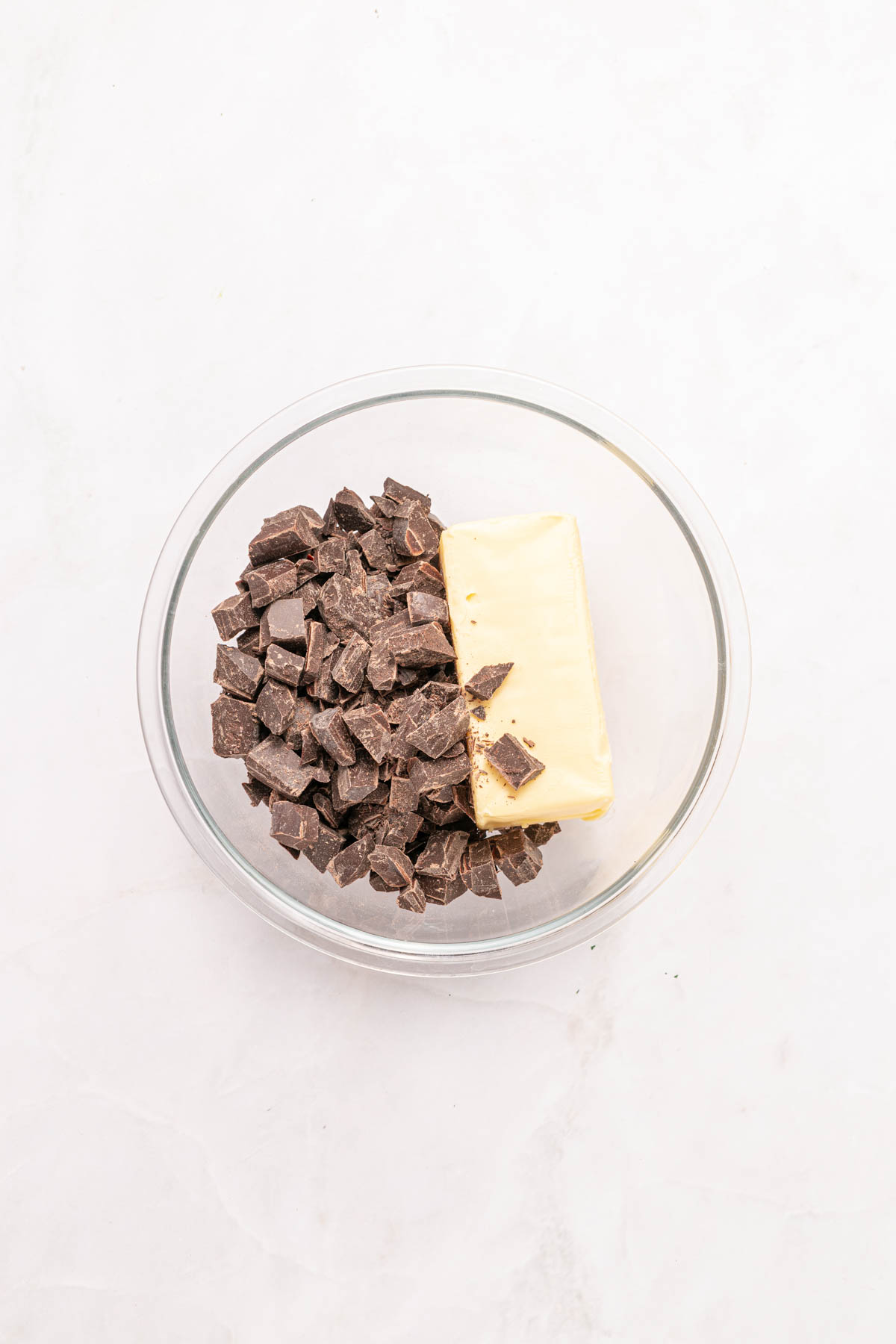 A glass bowl containing a stick of butter and chopped pieces of chocolate on a light surface.