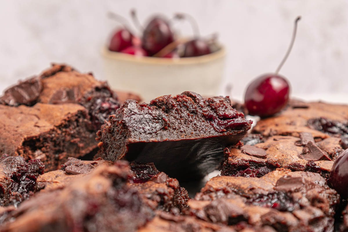 Close-up of chocolate cherry brownies with a moist, dense texture; fresh cherries and chunks of chocolate are visible, with a bowl of cherries in the background.