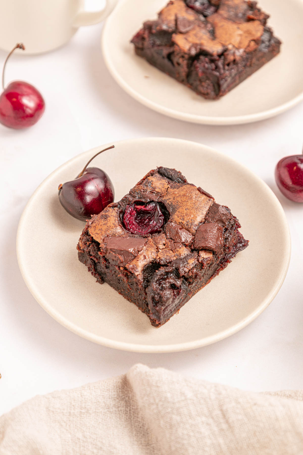 A square piece of chocolate brownie with a cherry on top sits on a white plate, alongside fresh cherries.