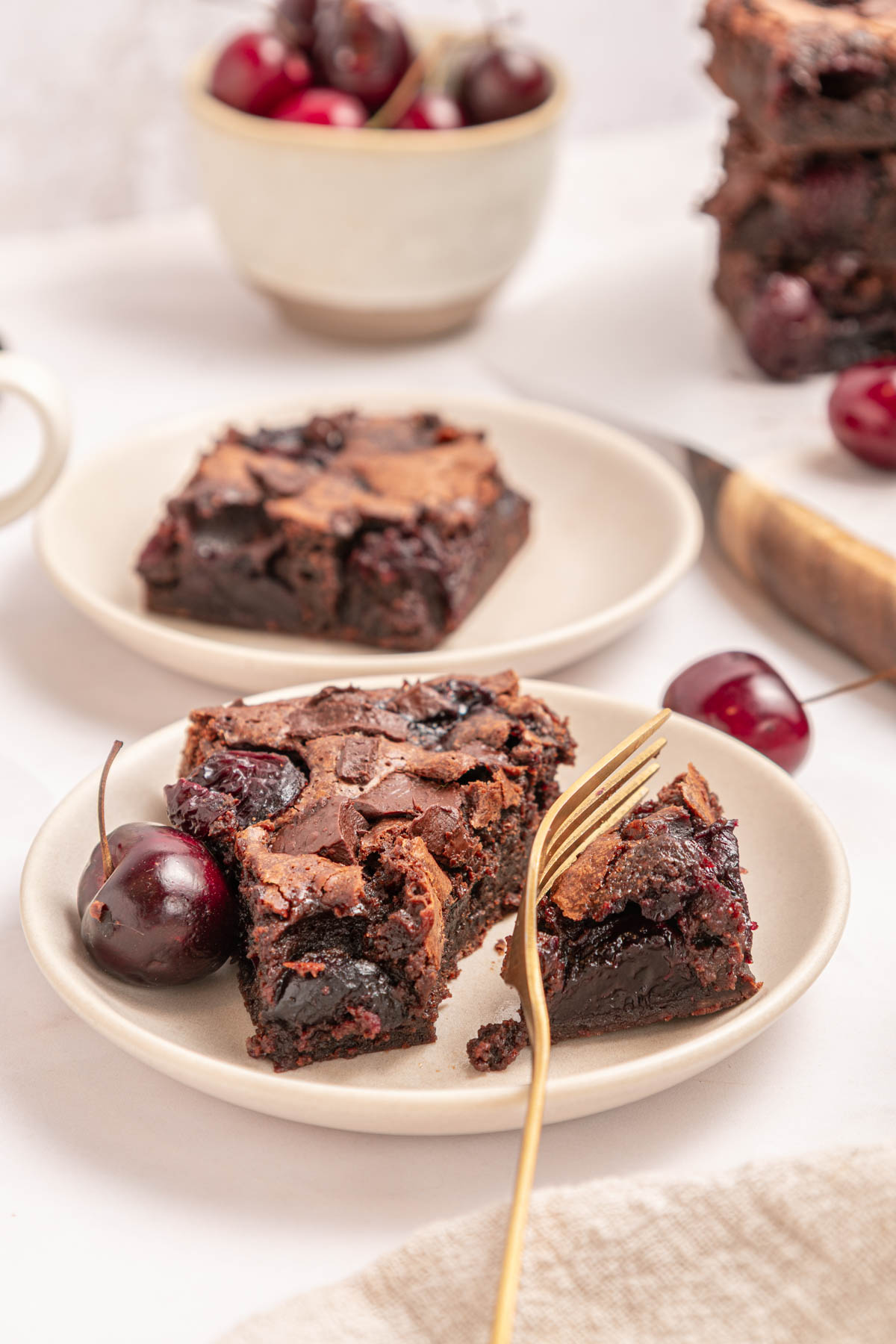 Two plates each hold a cherry chocolate brownie, with one brownie partially eaten and a gold fork beside it. Fresh cherries and stacked brownies are visible in the background.