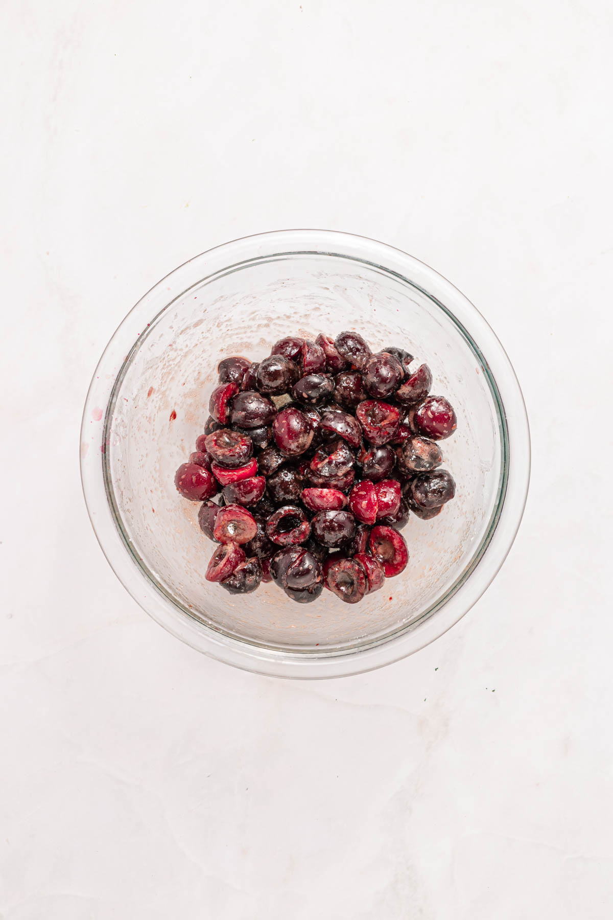 A glass bowl filled with halved dark cherries and other ingredients on a light-colored surface.