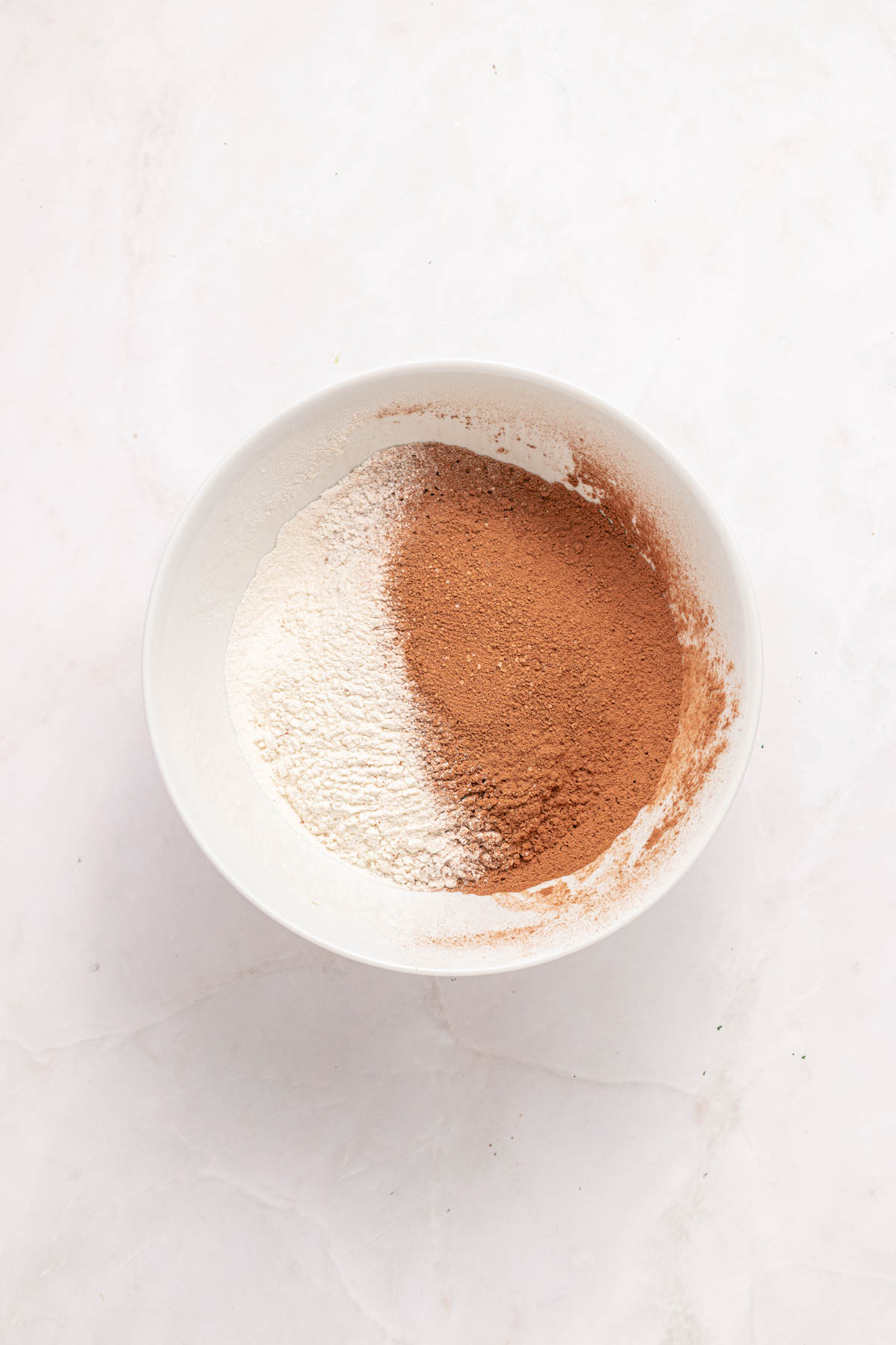 A white bowl containing flour and cocoa powder on a light marble surface.