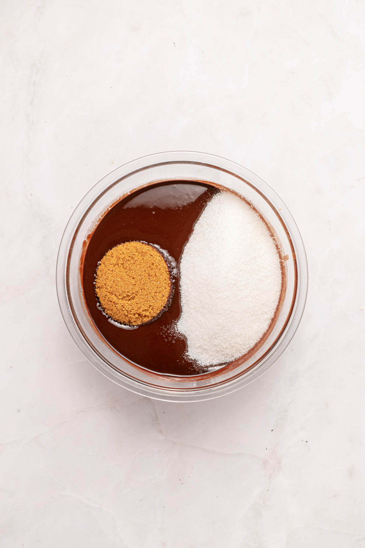 A glass bowl containing chocolate mixture, granulated sugar, and brown sugar on a white marble surface.