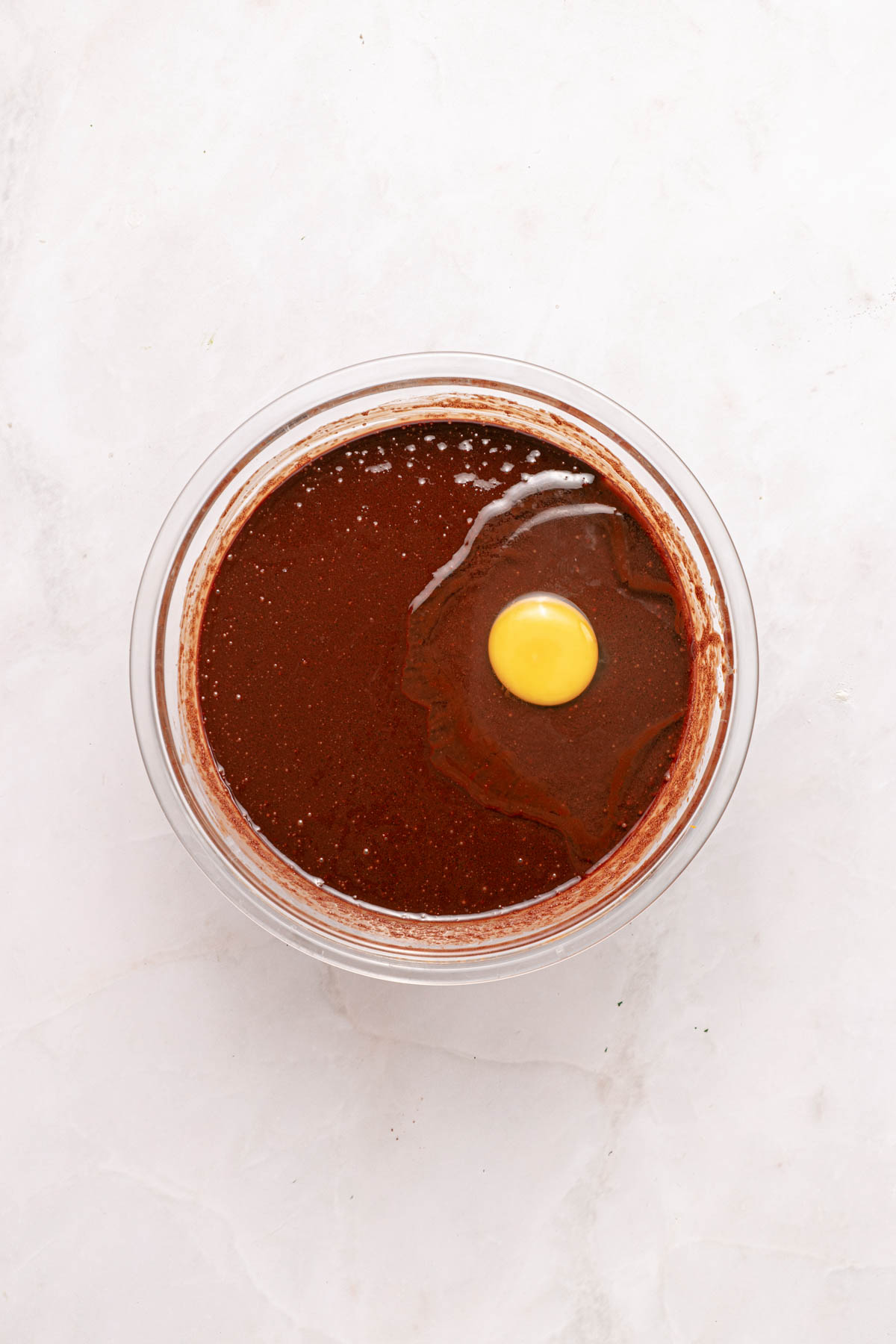A glass bowl containing chocolate batter with a raw egg cracked on top, viewed from above on a light surface.