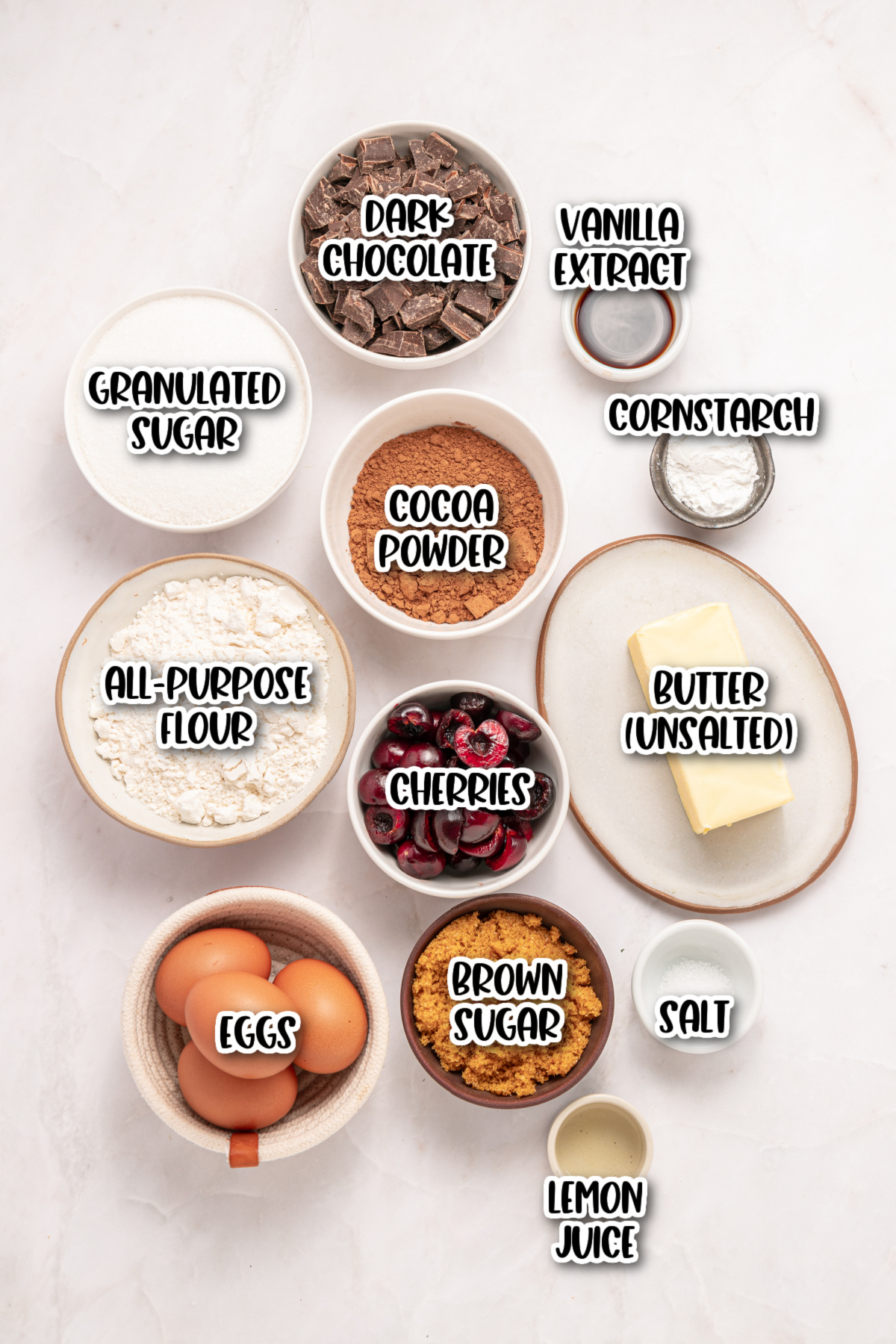 Top view of labeled baking ingredients including dark chocolate, vanilla extract, granulated sugar, cornstarch, cocoa powder, all-purpose flour, cherries, butter, eggs, brown sugar, salt, and lemon juice.