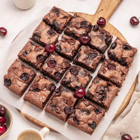 A batch of chocolate cherry brownies cut into squares on a round wooden board, garnished with fresh cherries.