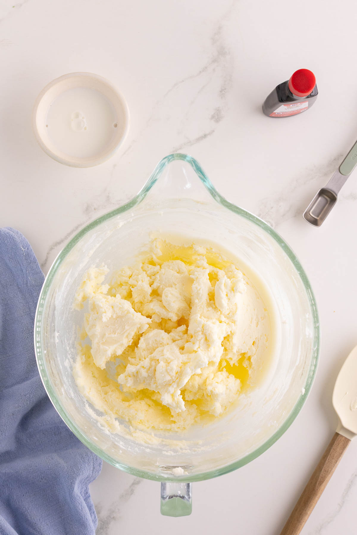 A glass mixing bowl with a yellow and white creamed mixture, surrounded by a measuring spoon, vanilla extract bottle, small bowl of milk, blue cloth, and spatula on a marble countertop.