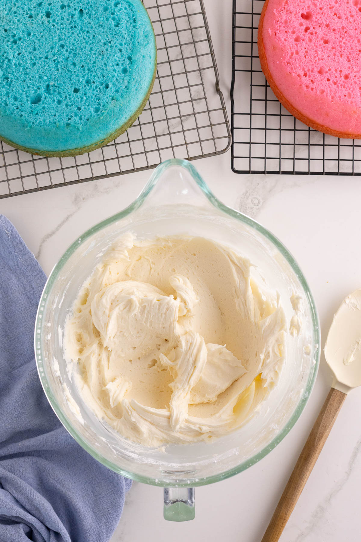A glass bowl of white frosting sits on a countertop next to a spatula, with blue and pink cake layers cooling on wire racks nearby.
