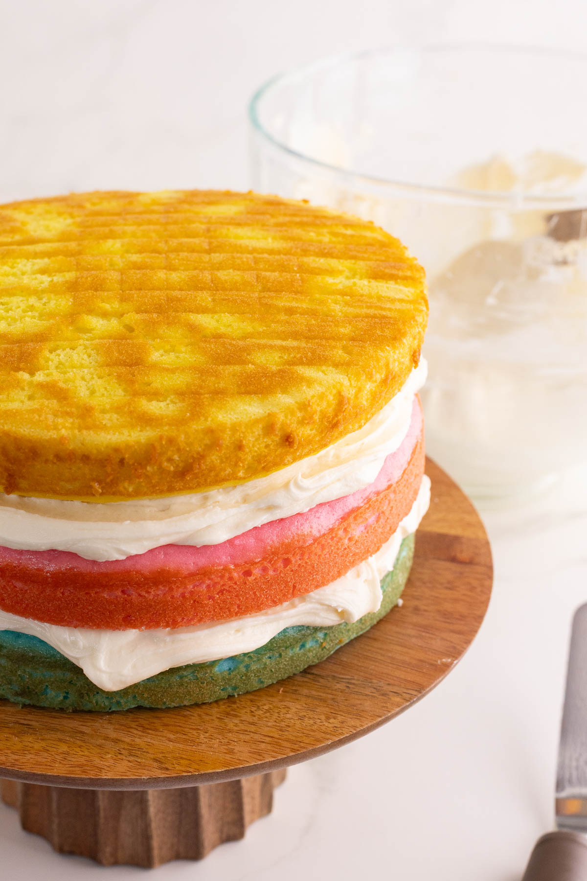 A partially frosted, three-layer cake with yellow, pink, and blue layers sits on a wooden cake stand. A glass bowl with frosting and a spatula is in the background.