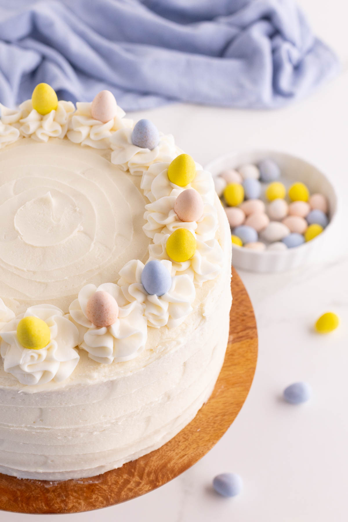 A round white frosted cake decorated with pastel candy eggs, placed on a wooden stand. A bowl of similar candy eggs and a light blue cloth are in the background.