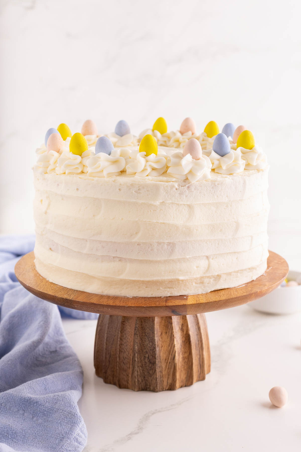 A round, white frosted cake decorated with small pastel candy eggs on top, displayed on a wooden cake stand.