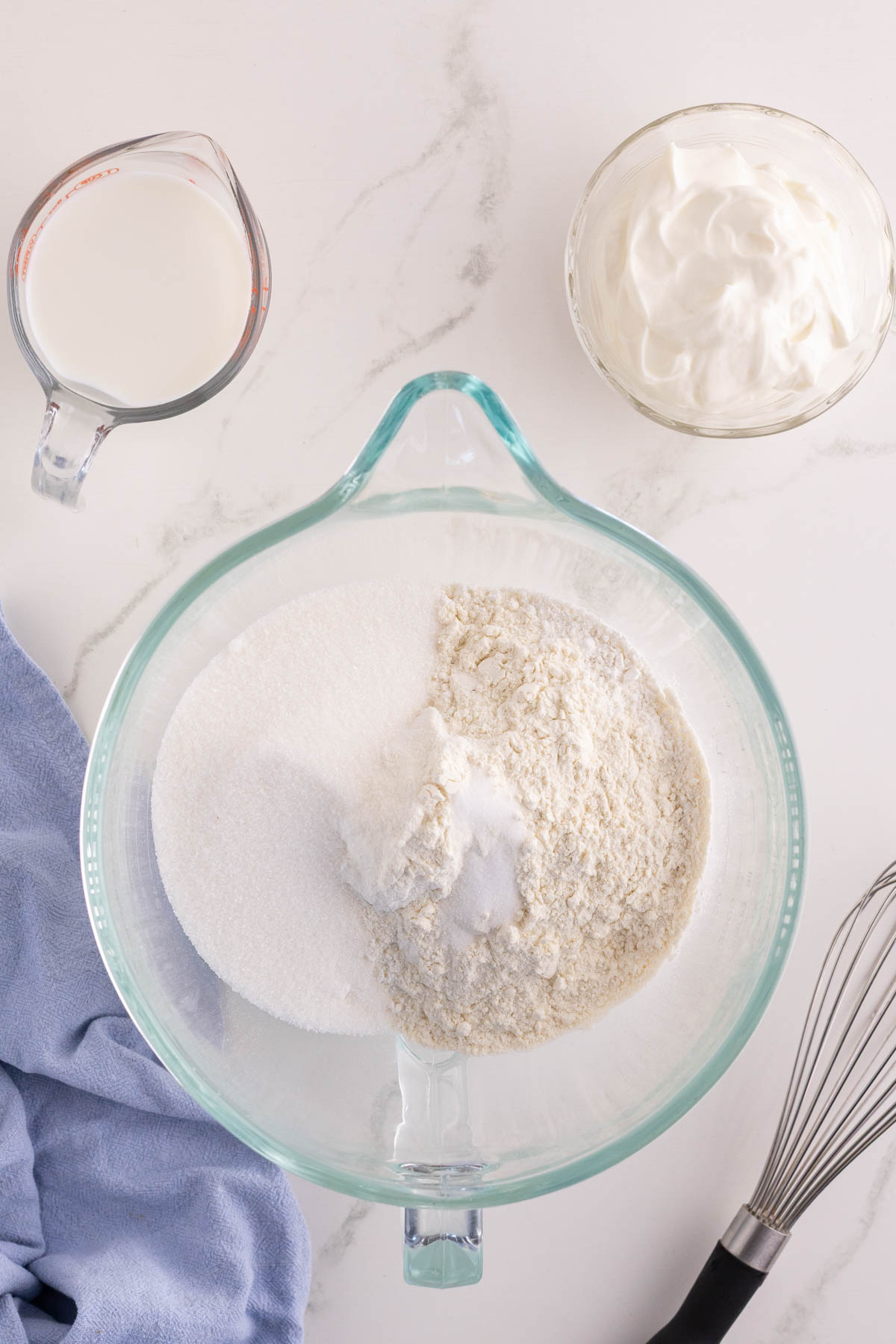 A glass bowl with flour and sugar, a cup of milk, a bowl of sour cream, a whisk, and a blue cloth on a white countertop.