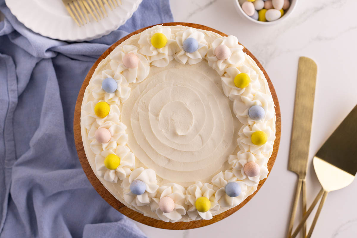 A round cake with white frosting, decorated with white rosettes and pastel-colored candy eggs on top, sits on a wooden cake stand. Nearby are serving utensils and a bowl of candy eggs.