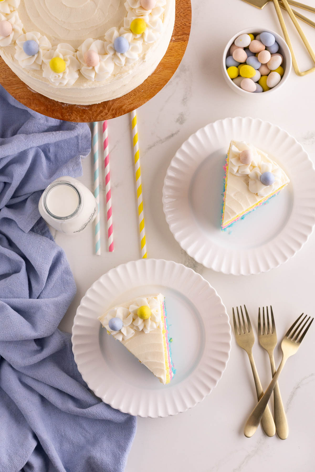 Two slices of frosted cake with pastel decorations are served on white plates, accompanied by forks, a small bottle of milk, and a bowl of candy on a marble surface.