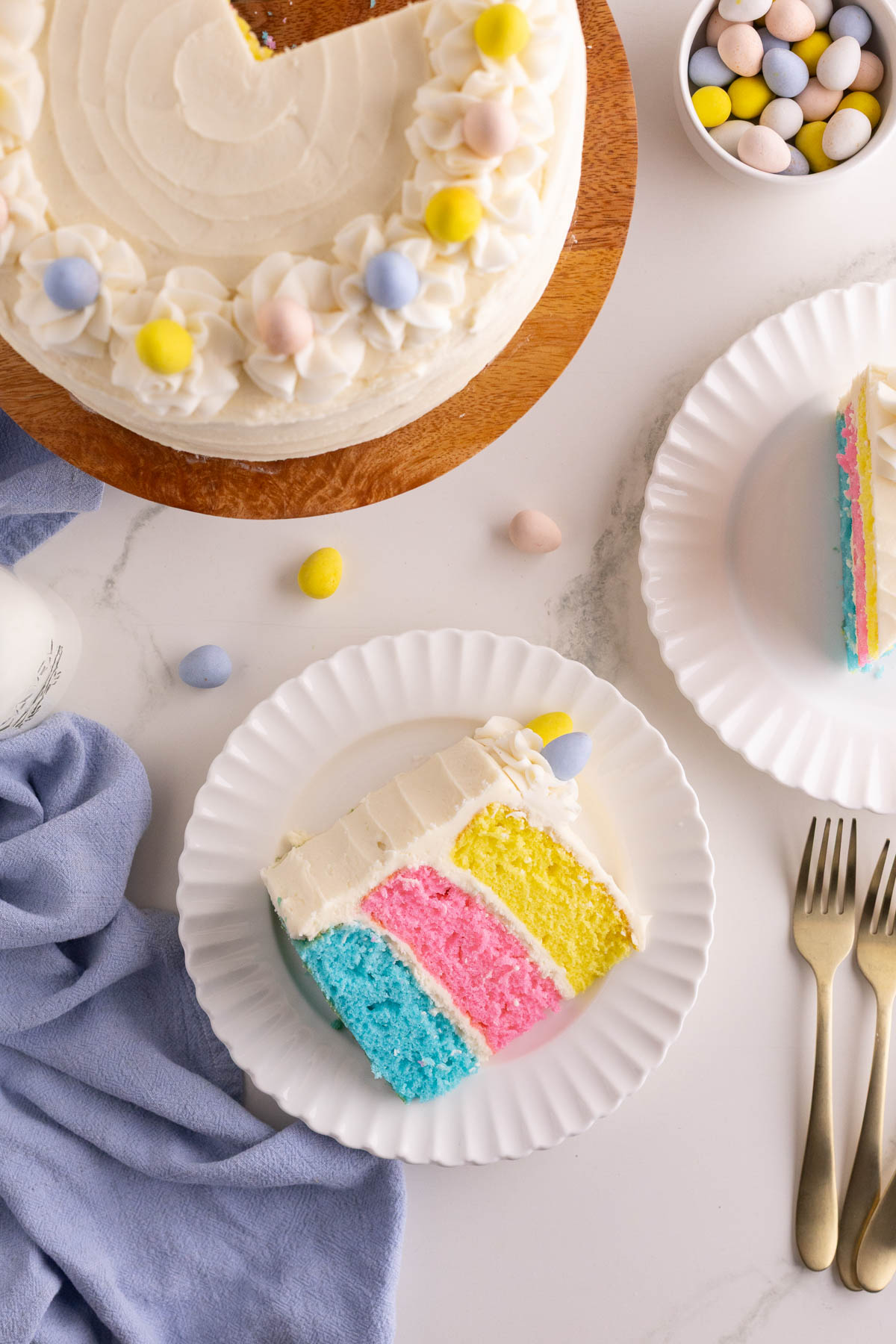 A slice of pastel-colored layer cake with white frosting on a plate, next to a whole cake, pastel candies, and two forks on a marble surface.