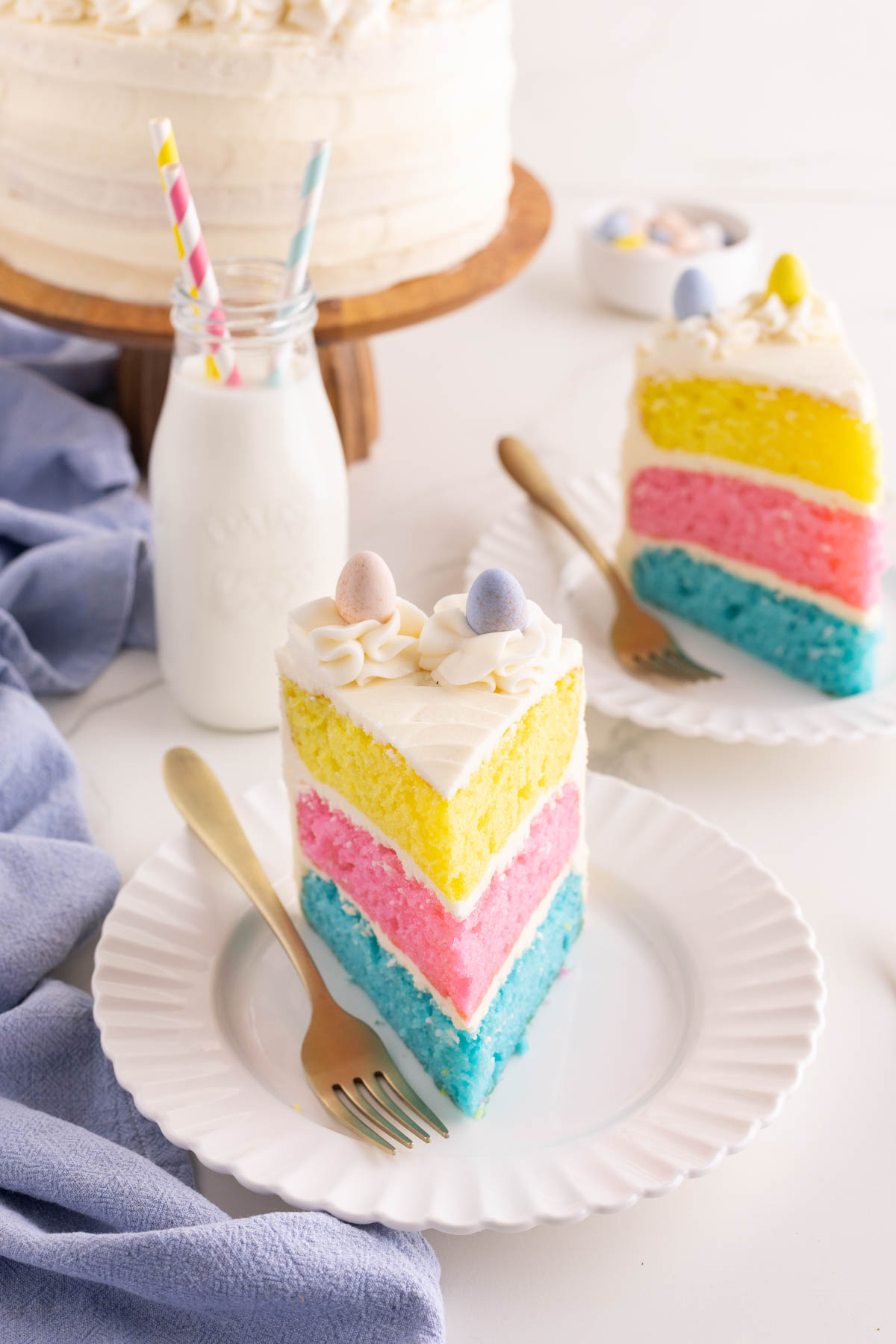 Two slices of three-layer pastel cake with white frosting and candy eggs on top, served on white plates with gold forks, next to a bottle of milk with striped straws.