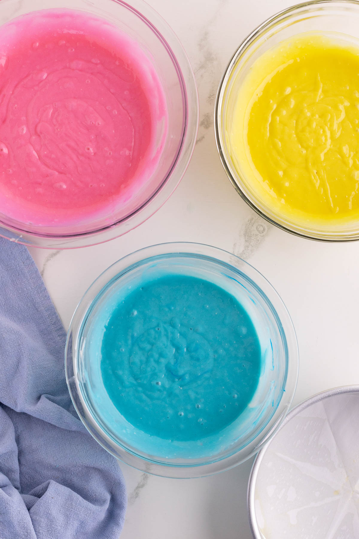 Three glass bowls filled with pink, yellow, and blue cake batter are arranged on a white surface next to a light purple cloth.
