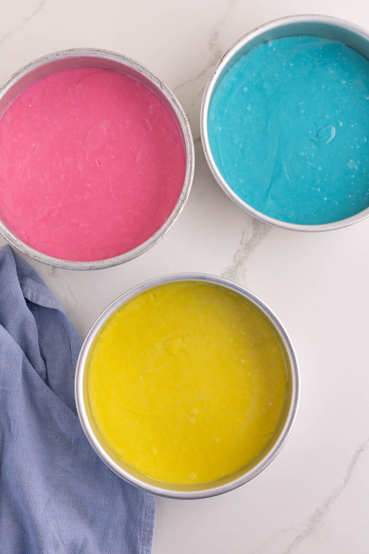 Three round cake pans filled with pink, blue, and yellow batter are placed on a white countertop next to a folded blue cloth.