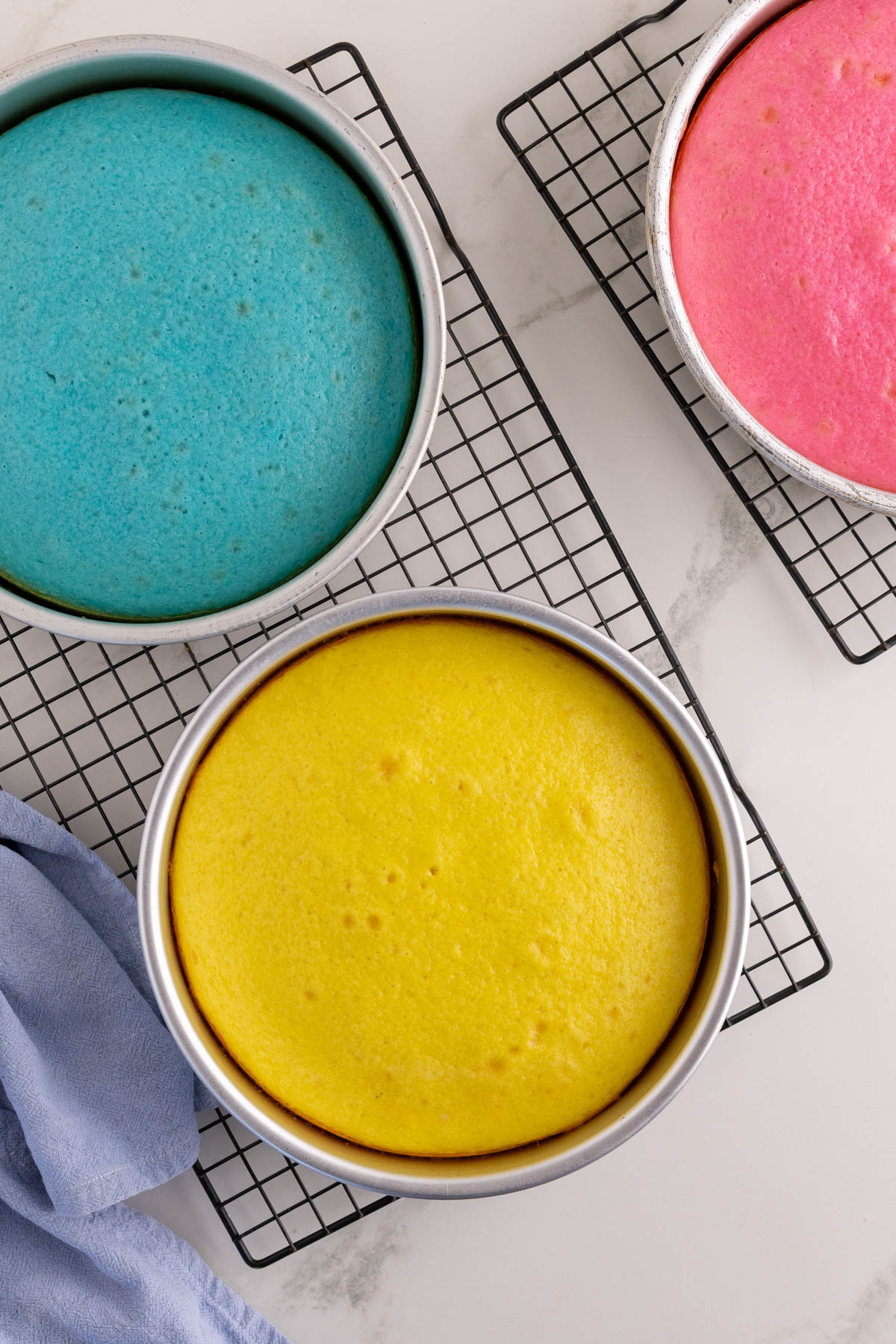 Three round cakes in baking pans, colored blue, yellow, and pink, are cooling on wire racks atop a white surface with a blue cloth nearby.