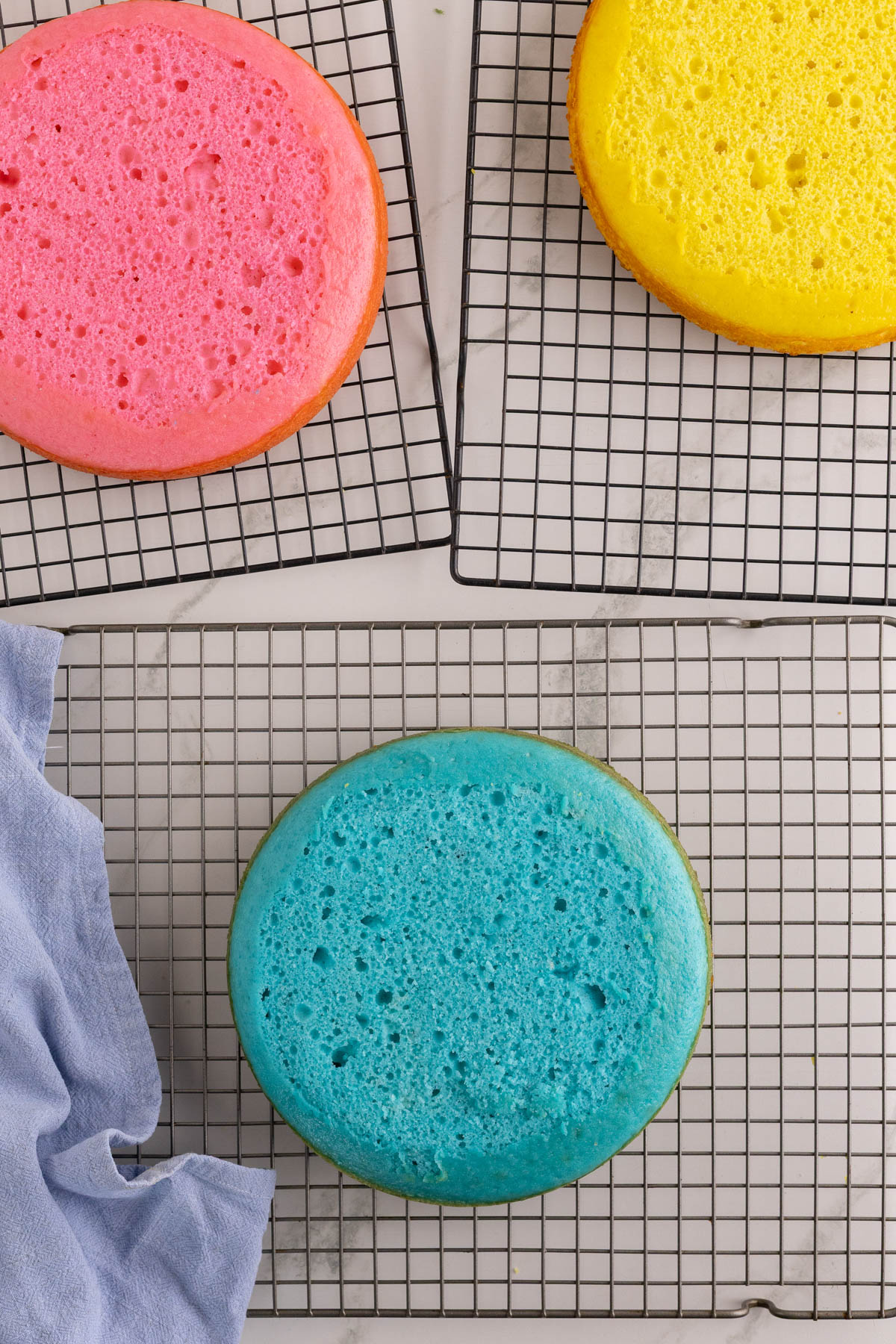 Three round cakes in pink, yellow, and blue colors are cooling on wire racks, with a light blue cloth partially visible on the left side.