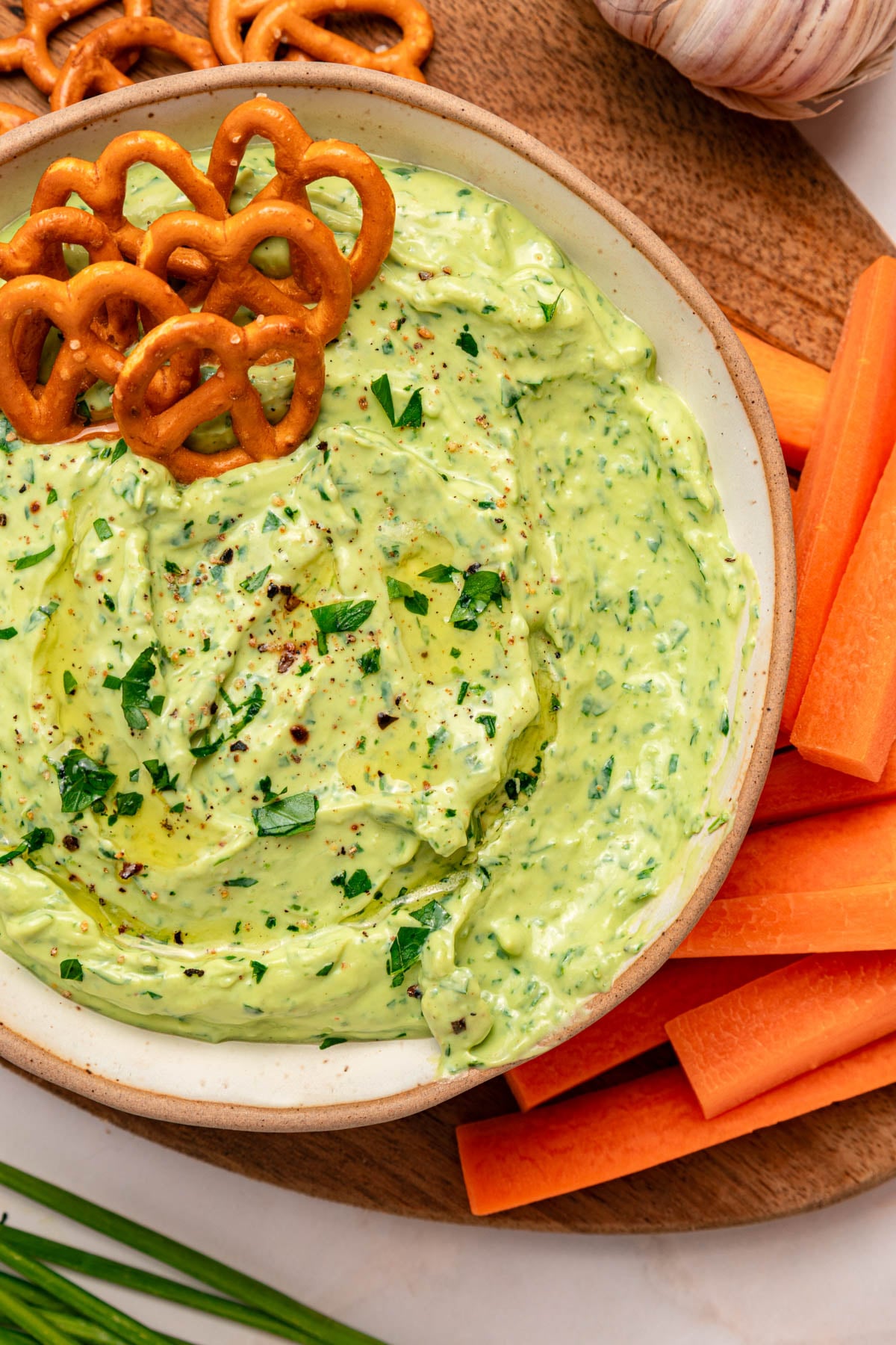 A bowl of green herb dip garnished with chopped herbs and pretzels, surrounded by carrot sticks on a wooden board.