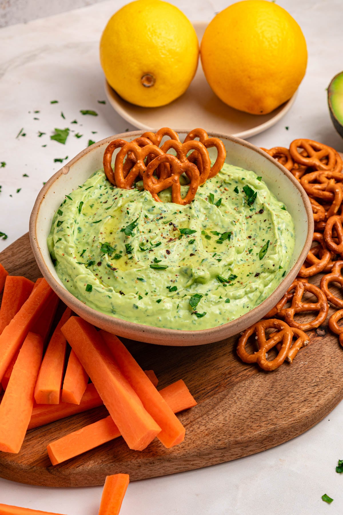 A bowl of creamy green dip garnished with chopped herbs, surrounded by carrot sticks and pretzels on a wooden board, with two lemons in the background.