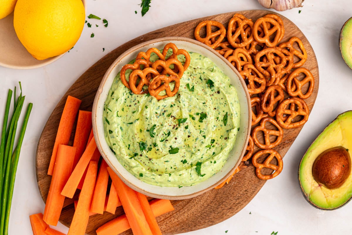 A bowl of creamy avocado dip garnished with herbs, surrounded by carrot sticks and pretzel twists on a wooden board.