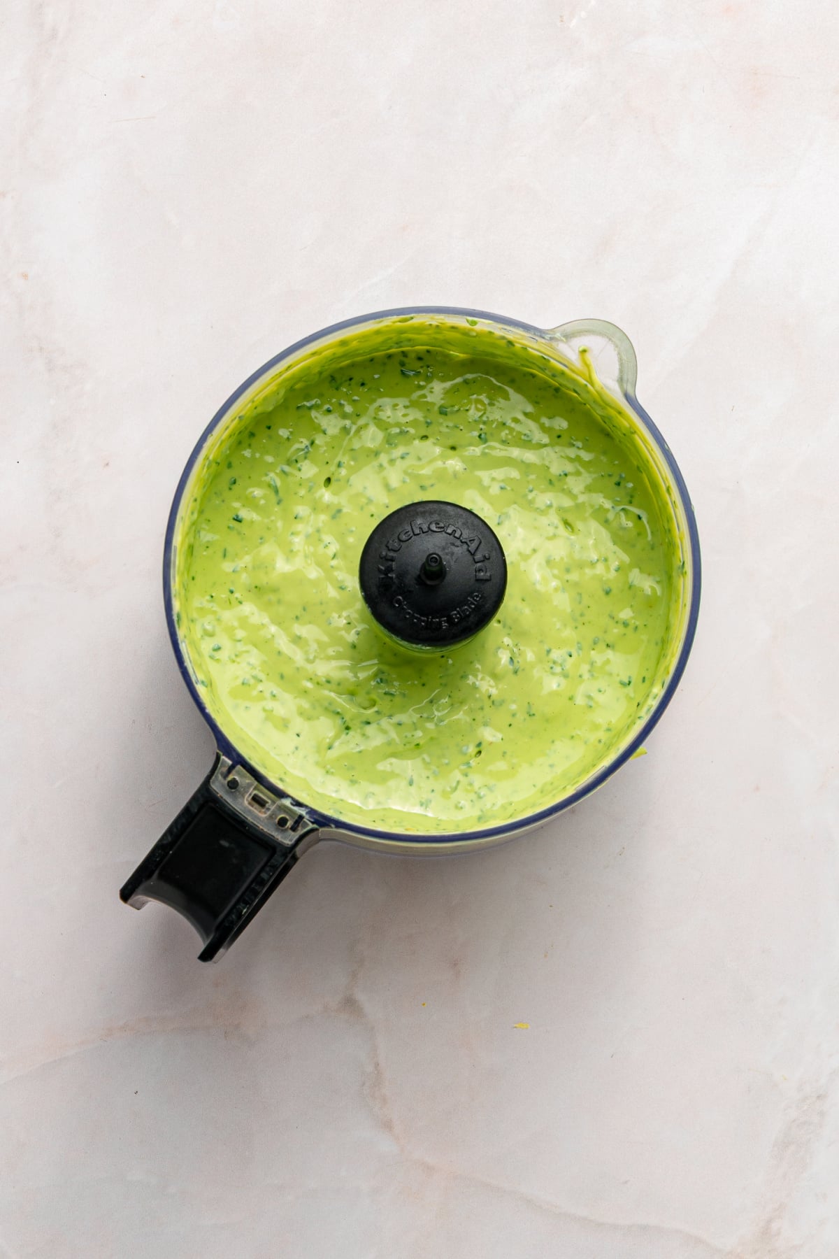 A food processor filled with a creamy green sauce or dressing, with visible herbs mixed in, sits on a light-colored surface.