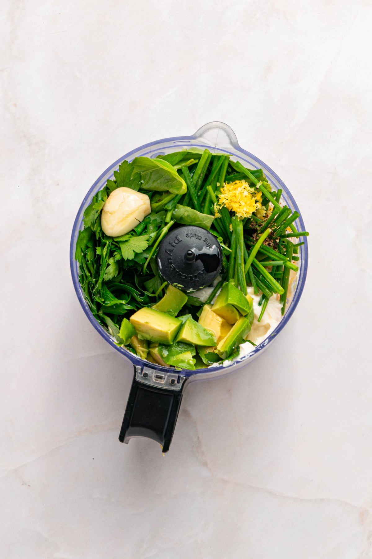 A food processor bowl containing fresh herbs, avocado, yogurt, lemon zest, and leafy greens on a light countertop.