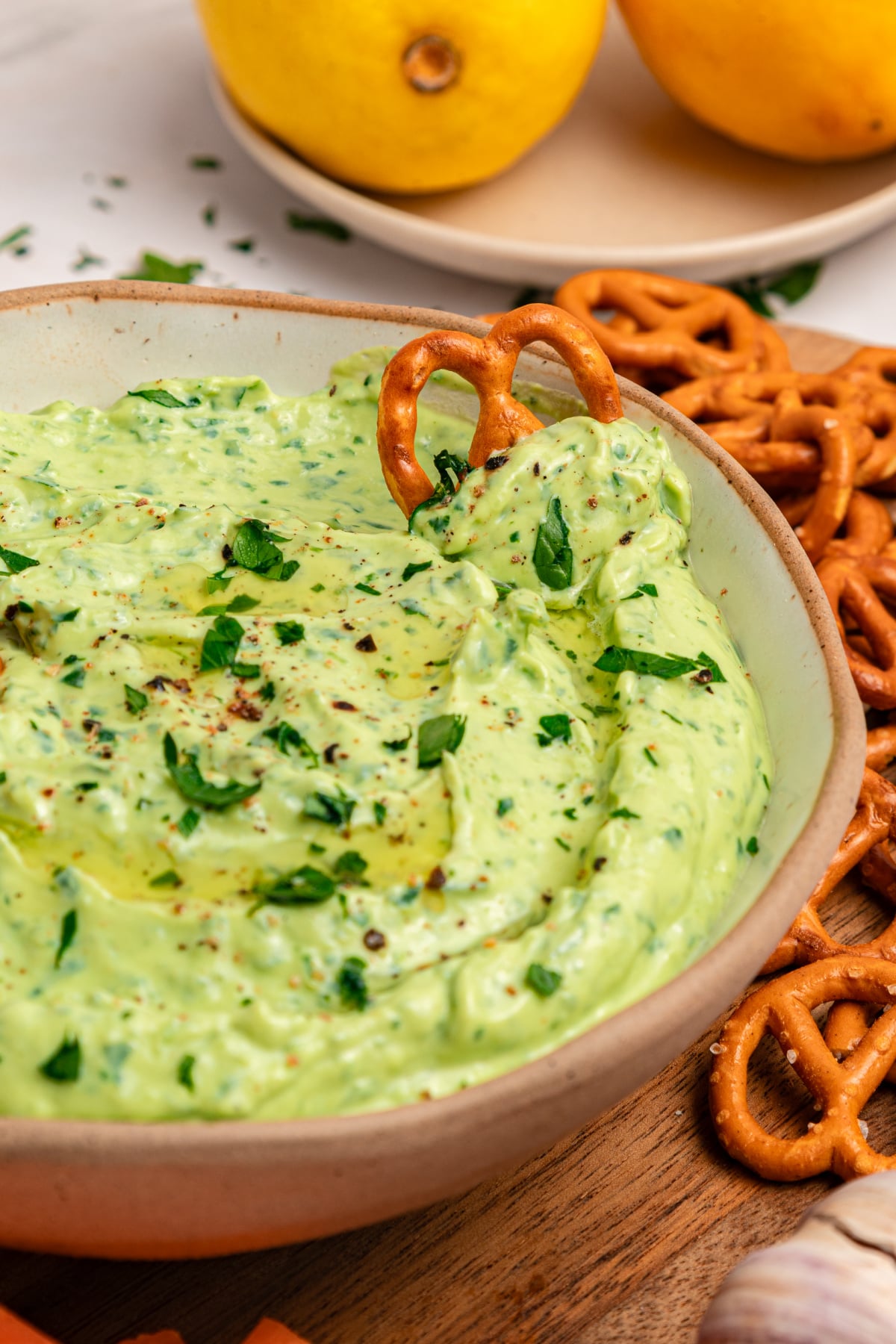A bowl of creamy green goddess dip garnished with chopped herbs, surrounded by pretzels, with lemons in the background.