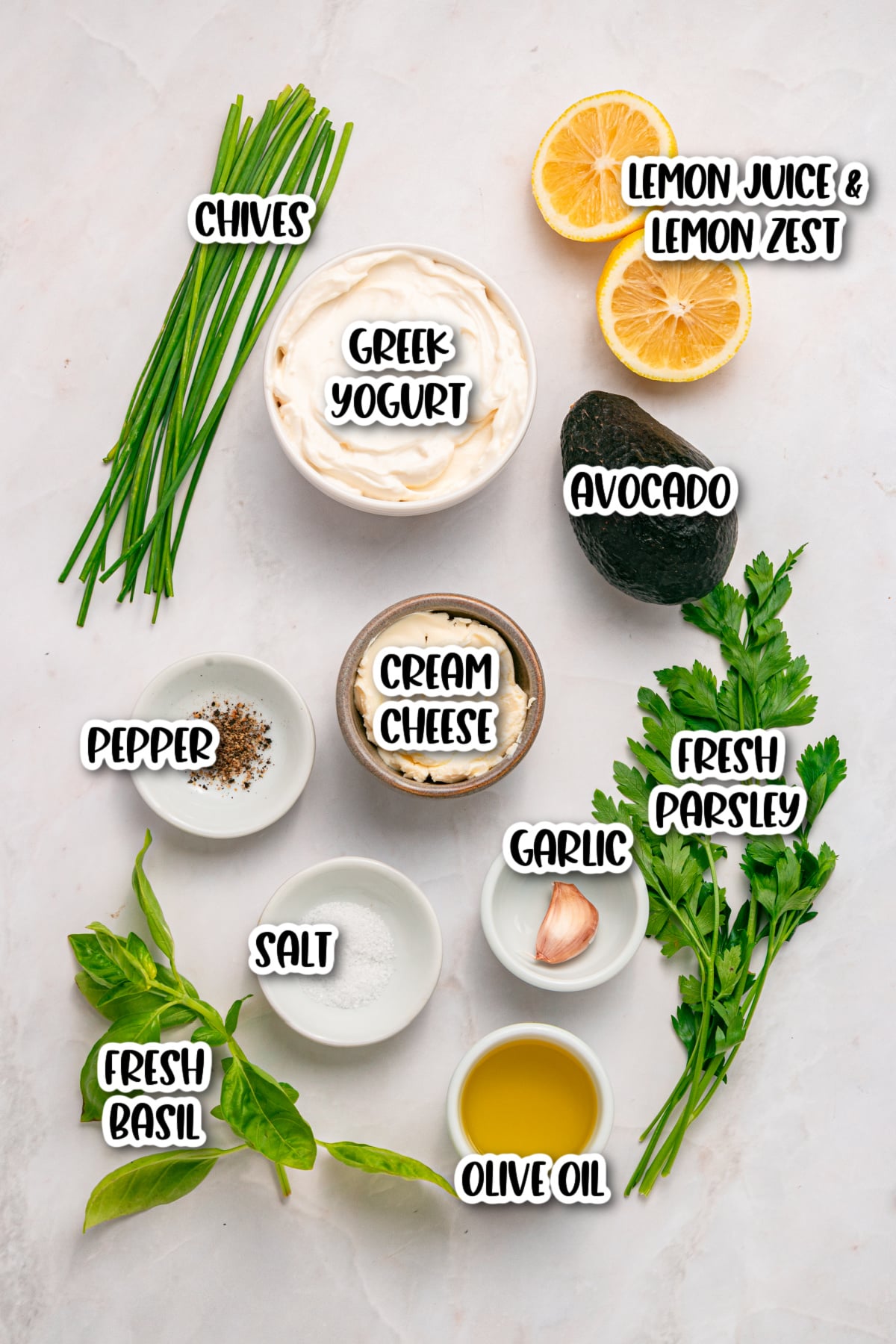 A flat lay of ingredients including chives, Greek yogurt, lemon, avocado, fresh parsley, fresh basil, cream cheese, garlic, olive oil, salt, and pepper on a white background.