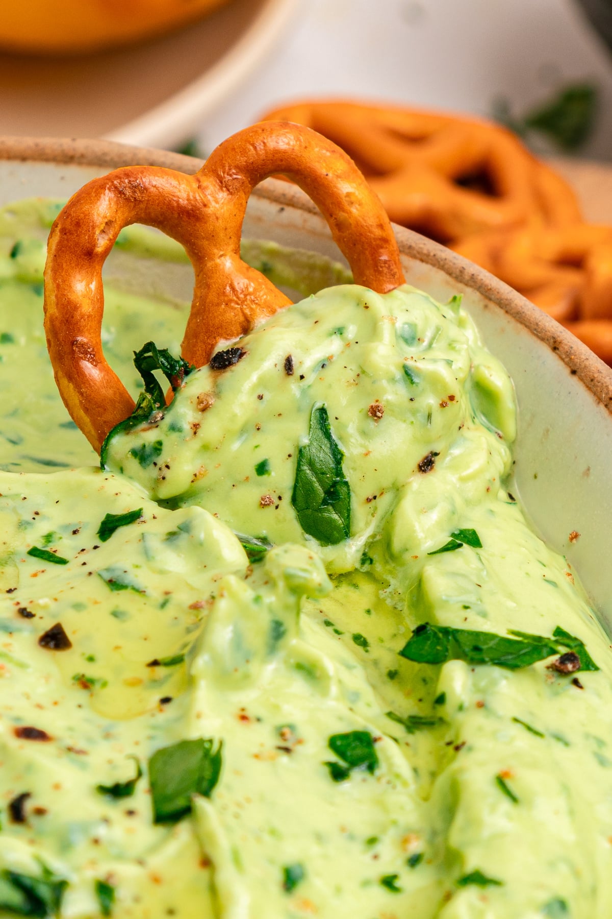 A pretzel is dipped into a creamy green herb dip, garnished with chopped herbs and cracked black pepper.