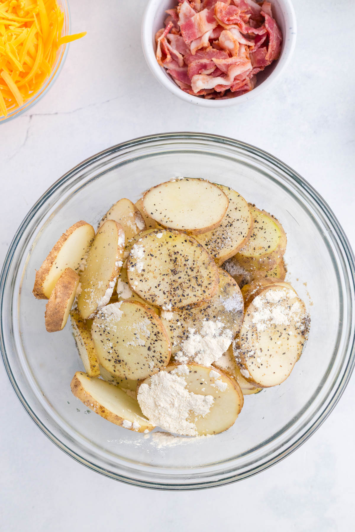 A glass bowl of sliced potatoes coated with flour and black pepper sits beside bowls of shredded cheese and cooked bacon on a white surface.