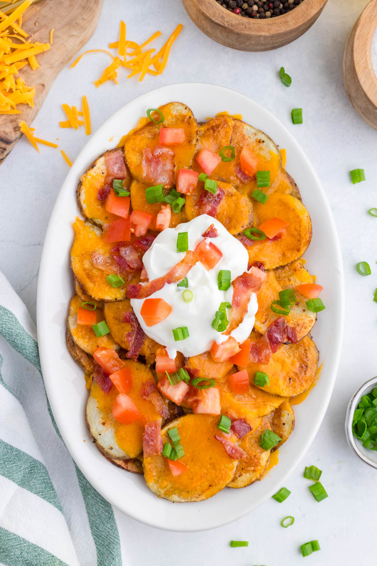 A white oval dish filled with sliced potatoes topped with melted cheddar cheese, bacon pieces, chopped tomatoes, green onions, and a dollop of sour cream.