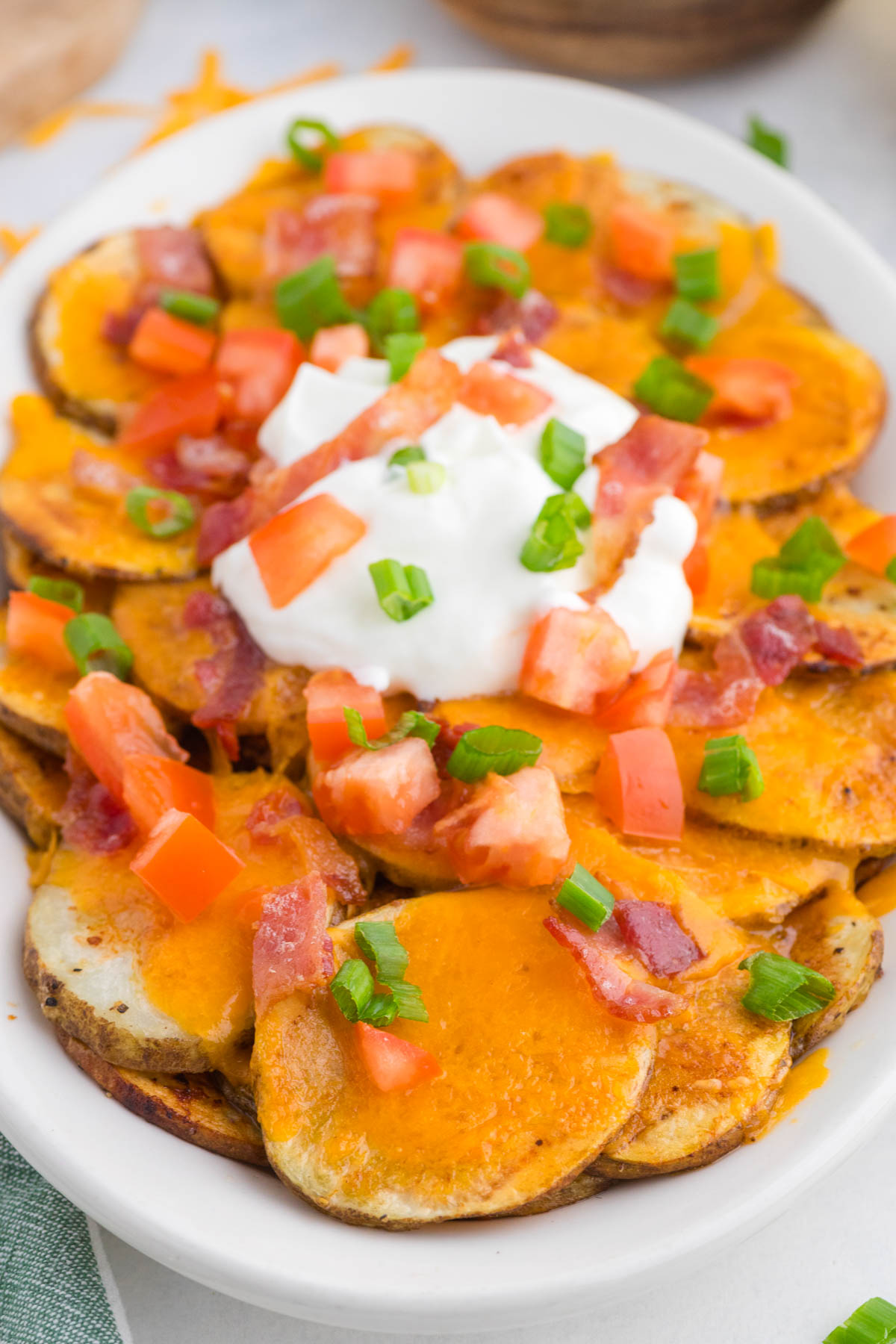 A plate of sliced potatoes topped with melted cheddar cheese, diced tomatoes, bacon bits, chopped green onions, and a dollop of sour cream.
