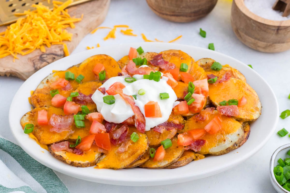 A plate of sliced baked potatoes topped with melted cheddar cheese, bacon, chopped tomatoes, green onions, and a dollop of sour cream.