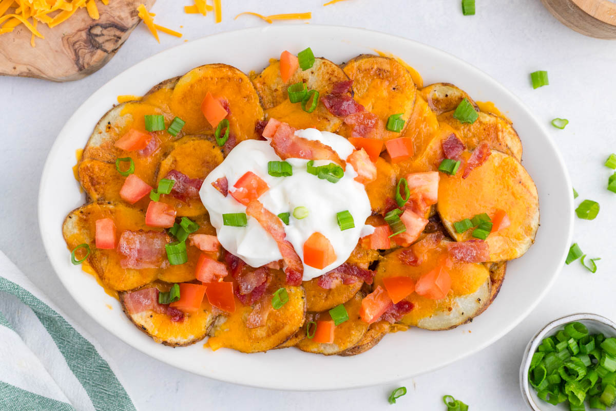 Oval plate of sliced potatoes topped with melted cheddar cheese, bacon pieces, chopped tomatoes, green onions, and a dollop of sour cream in the center.