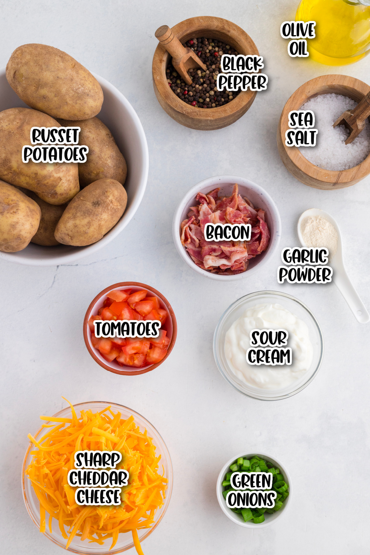 Top-down view of ingredients for Irish nachos, including russet potatoes, bacon, tomatoes, sharp cheddar cheese, sour cream, green onions, black pepper, sea salt, olive oil, and garlic powder.