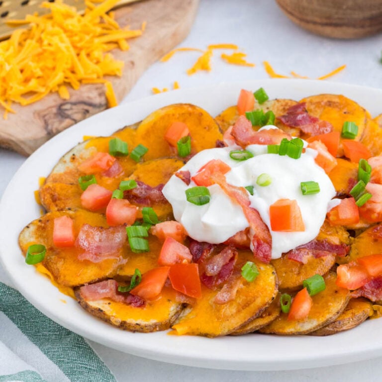 A plate of potato slices topped with melted cheddar cheese, bacon, chopped tomatoes, green onions, and a dollop of sour cream. Shredded cheese is visible in the background.