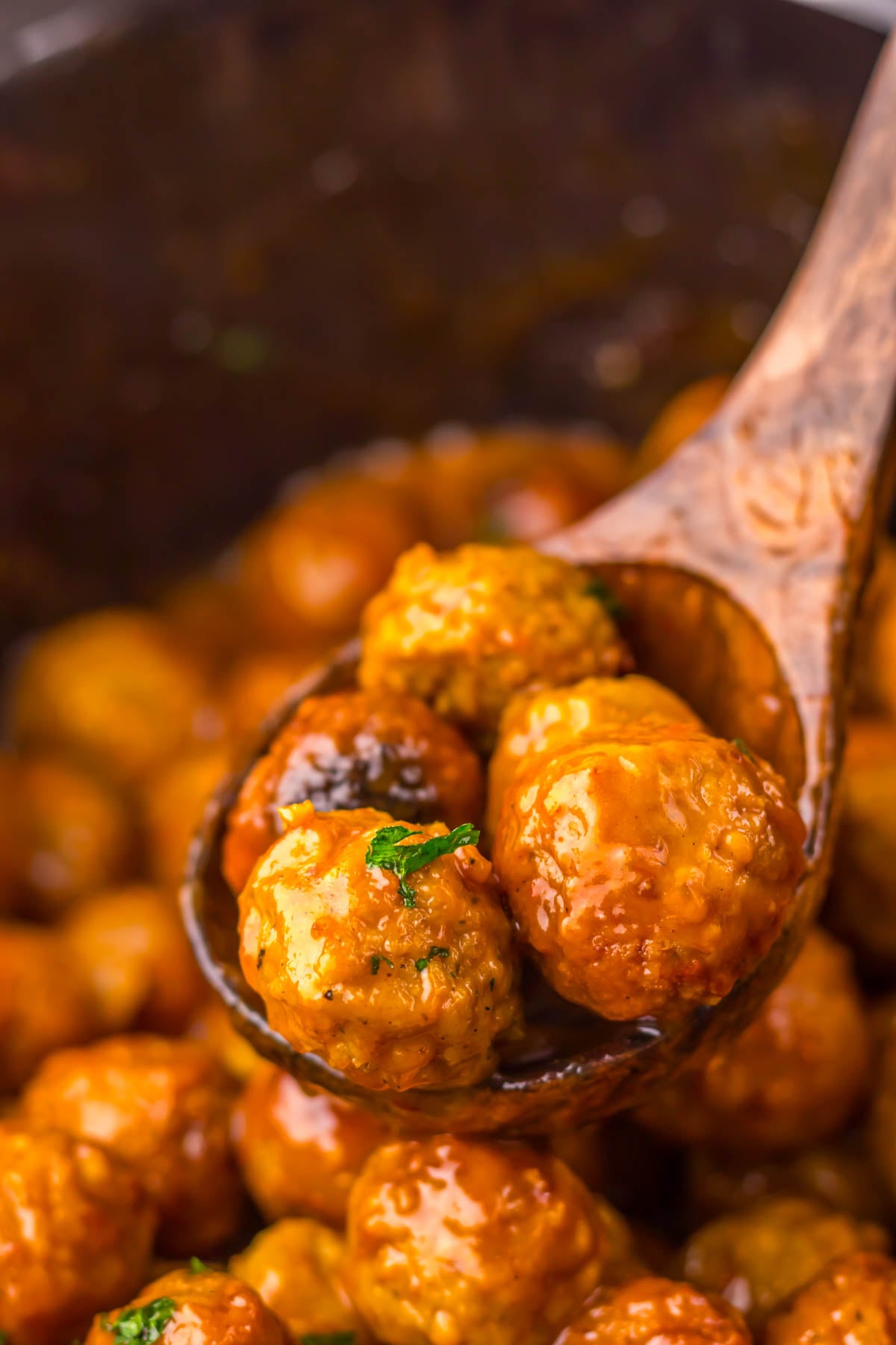 A wooden spoon holds several glazed meatballs garnished with a small sprig of parsley, with more meatballs visible in the background.