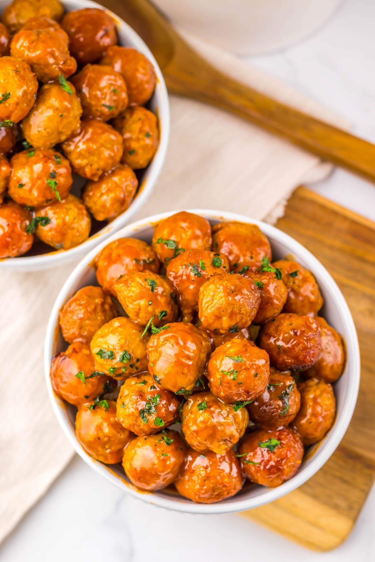 Two white bowls filled with glazed meatballs topped with chopped herbs are placed on a wooden cutting board and a white cloth.