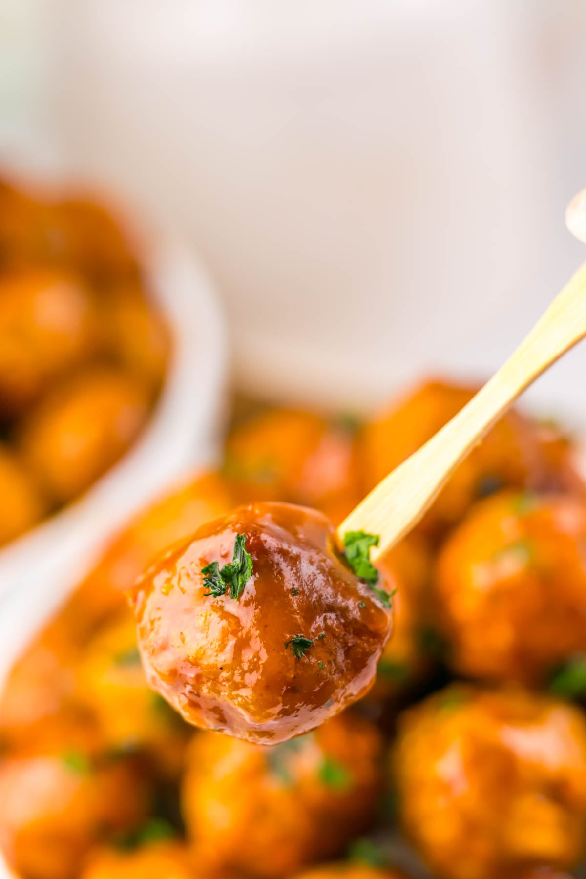 A close-up of a glazed meatball garnished with herbs, held on a wooden skewer, with more meatballs blurred in the background.