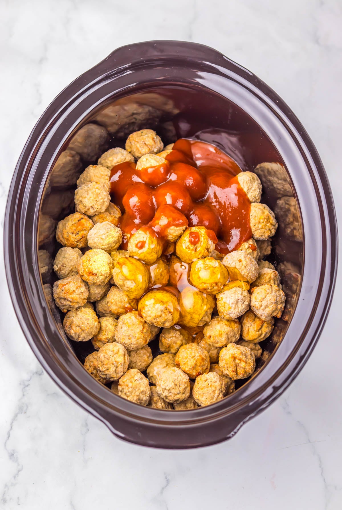 A slow cooker filled with frozen meatballs topped with ketchup and grape jelly on a marble countertop.