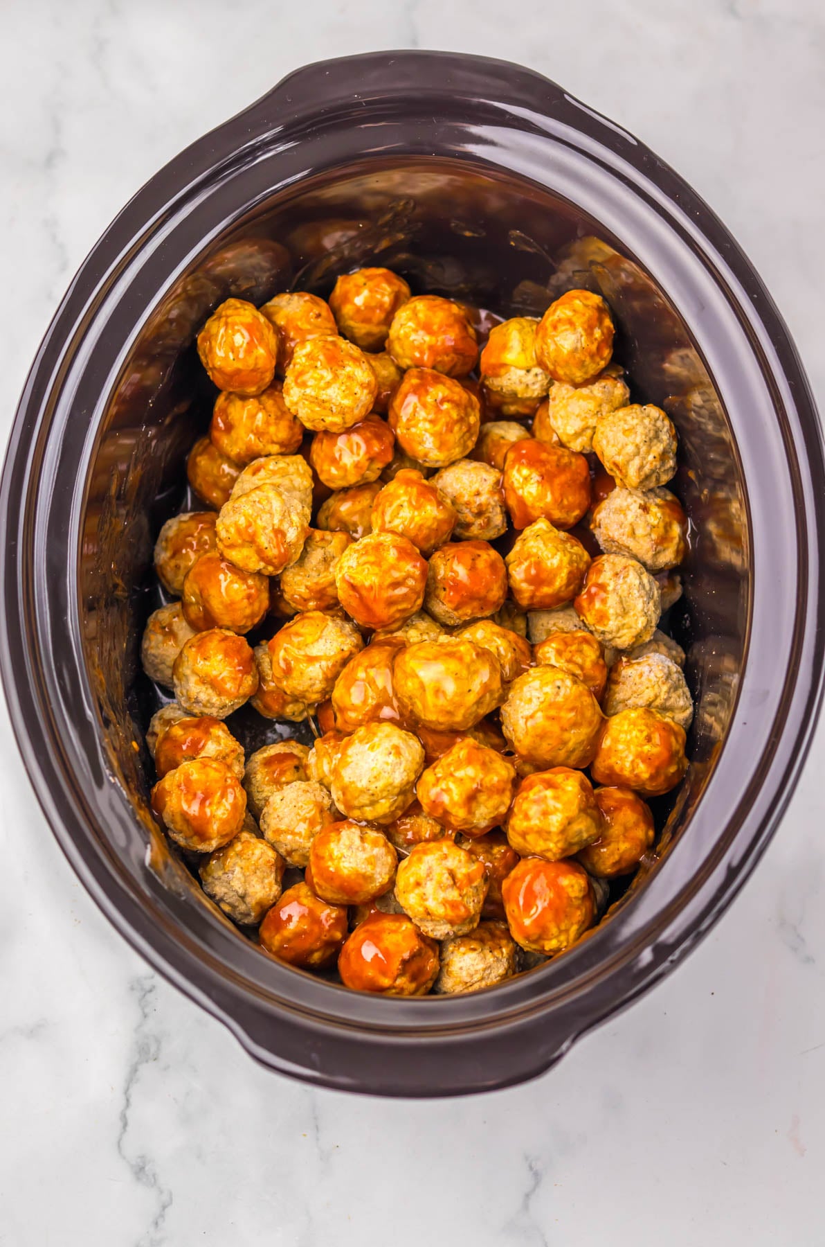A slow cooker filled with cooked meatballs coated in a reddish-orange sauce, placed on a light-colored countertop.
