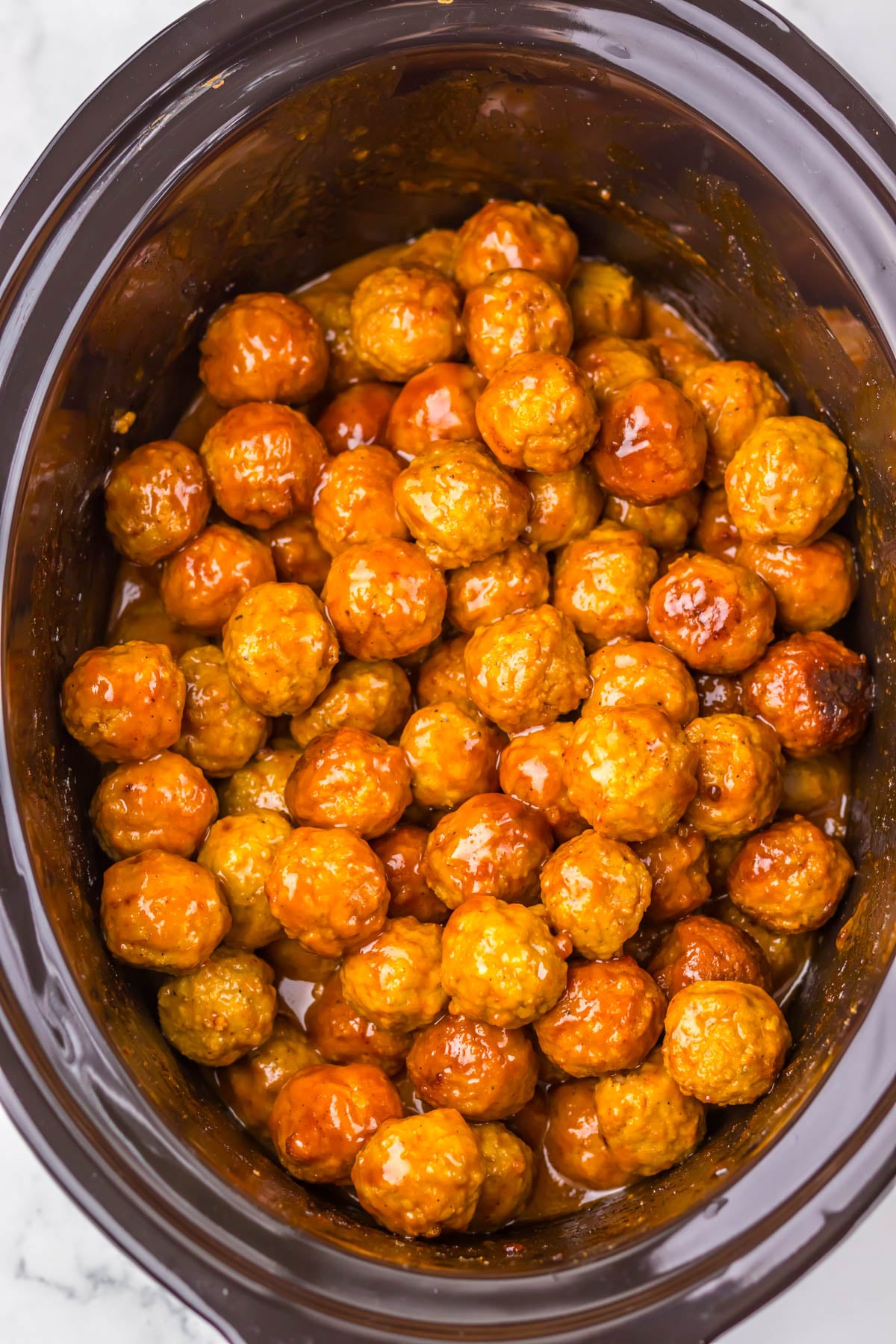 An overhead view of cooked meatballs coated in orange sauce inside a black slow cooker.